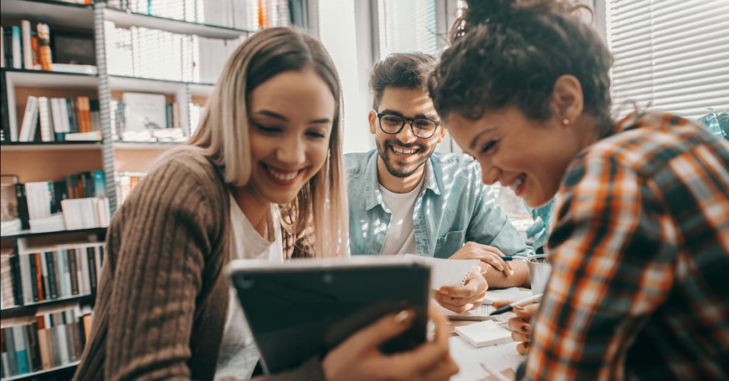 Three happy students dressed casual using tablet for school project and sitting at desk in library.